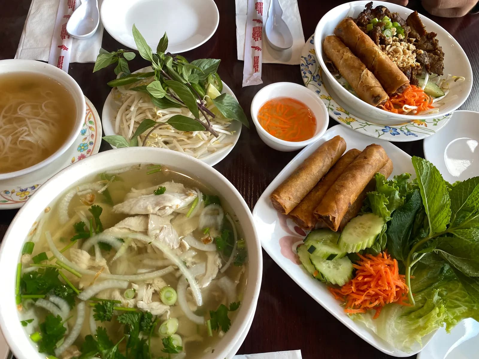 A Vietnamese family dinner spread: chicken pho, spring rolls, grilled pork over vermicelli and a plate of fresh herbs